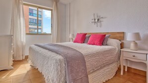 A neatly arranged bedroom with a large bed, two bedside tables, a nightstand, a window with curtains, and a view of a building outside. - Cosy apartment for 4 people with pool, A/C, WIFI, TV and terrace (Benissa)