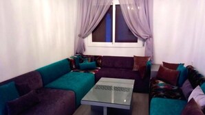 A living room with a glass coffee table, purple and teal sofas, and a window with curtains.