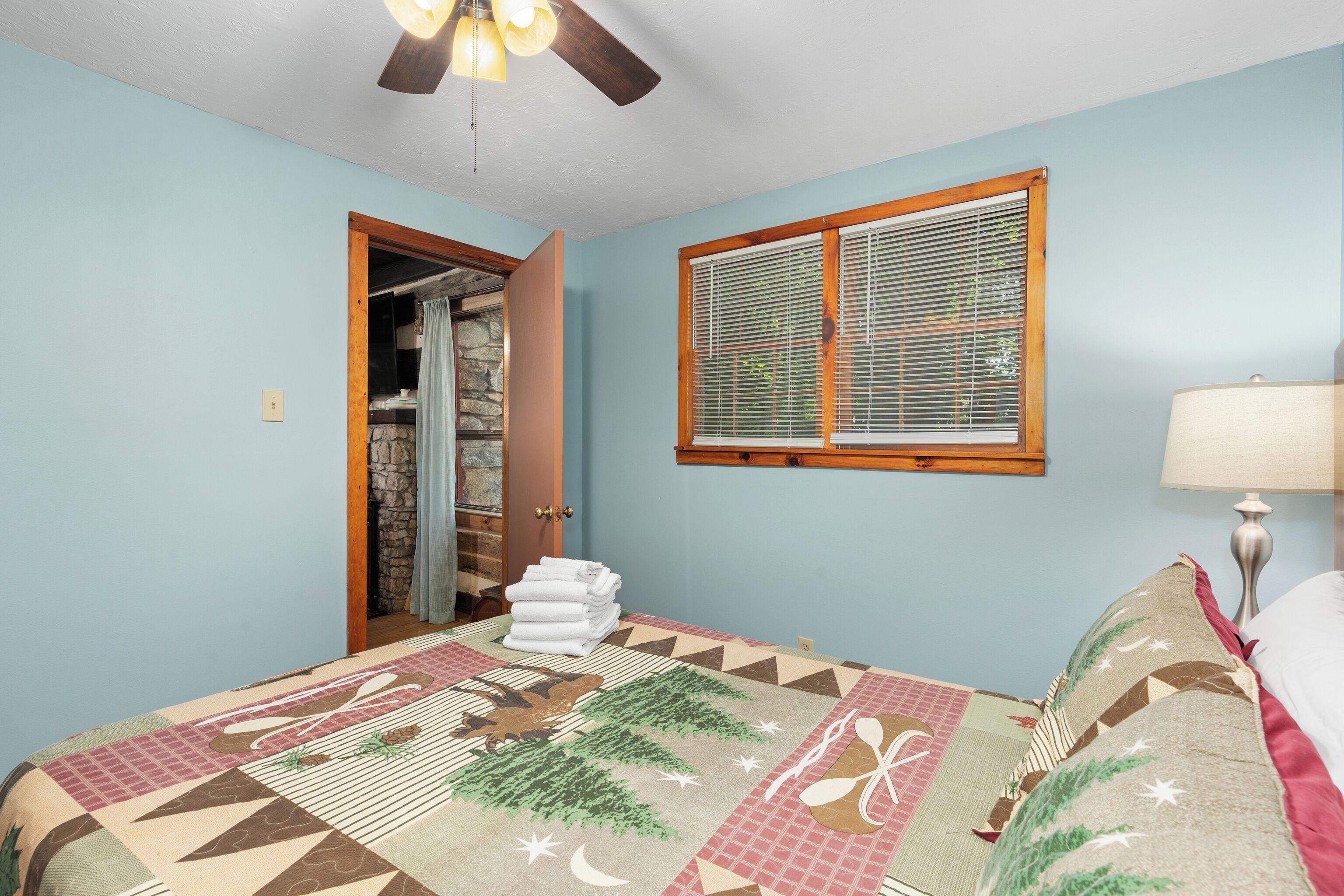 A bedroom with a bed, a window with blinds, a ceiling fan, and a door leading to a bathroom.