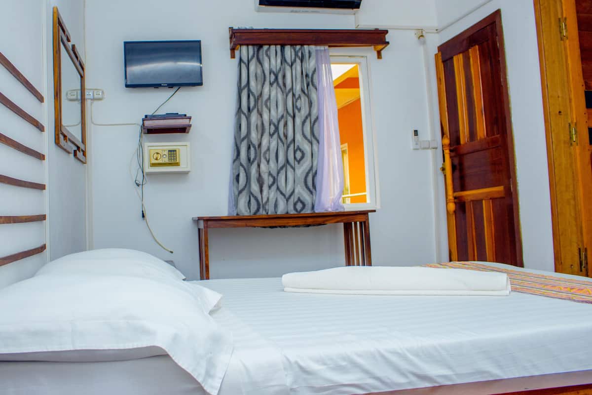 A single-bed room with a wooden headboard, a television mounted on the wall, a bedside table with a lamp, and a patterned curtain.