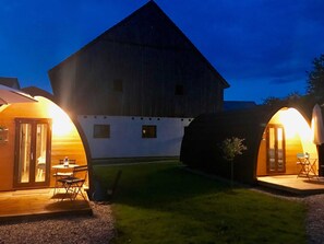 Un bâtiment de style grange, doté d’une grande porte et de grandes fenêtres, d’un espace extérieur couvert avec une table et des chaises, et d’une cour gazonnée.