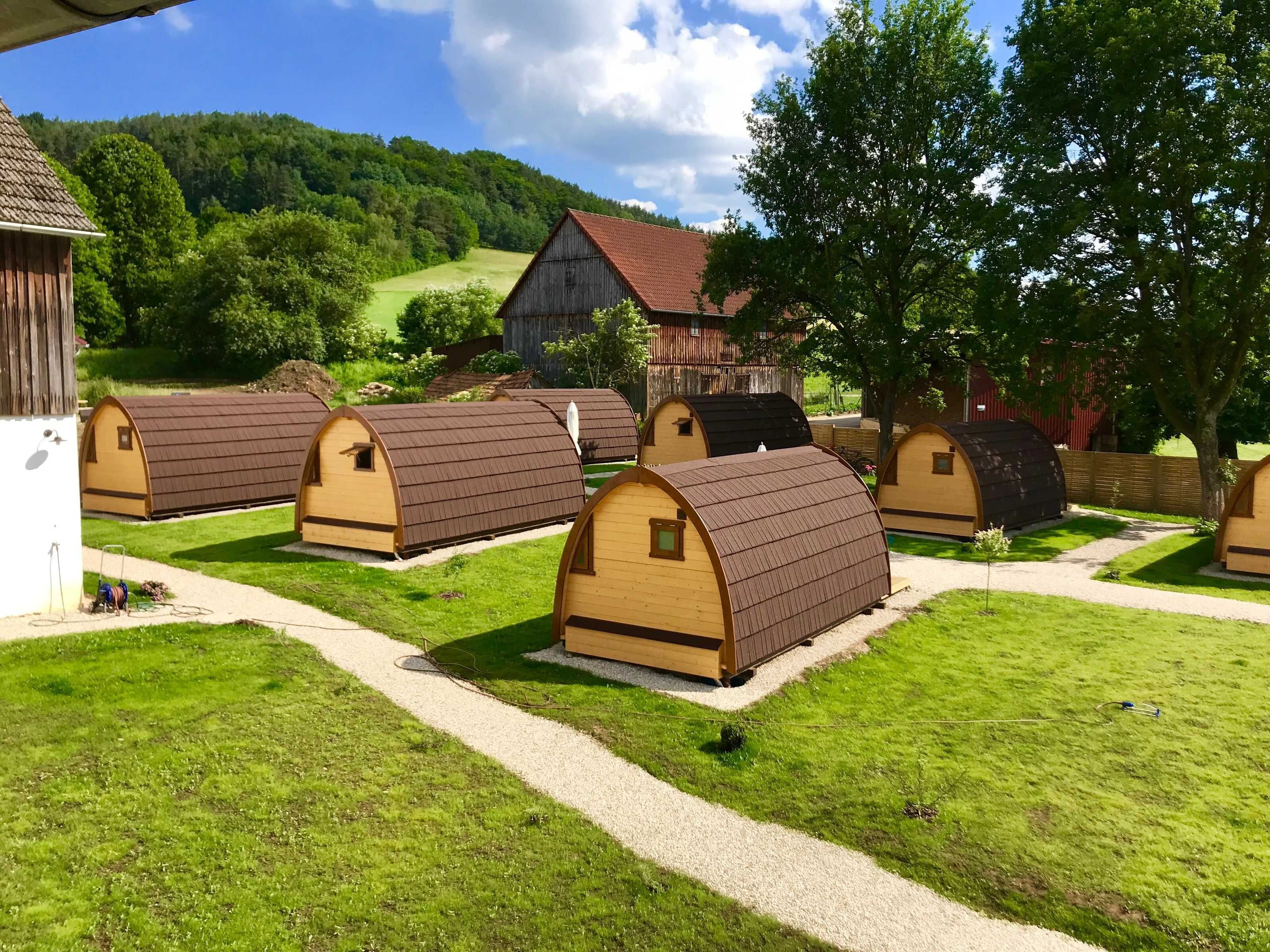 A row of small, wooden, arched-roof cabins with gravel paths between them, set in a rural landscape with a house in the background.