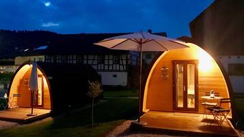 Deux cabanes en bois en forme de dôme, avec une table et des chaises à l’extérieur, un grand parasol et une maison en arrière-plan.