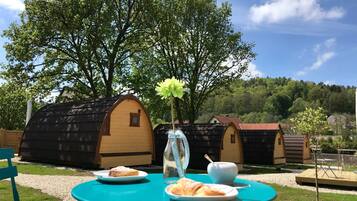 Une table turquoise sur laquelle se trouve une assiette de nourriture, un vase en verre avec une fleur et une tasse. On aperçoit des bâtiments en bois au fond.