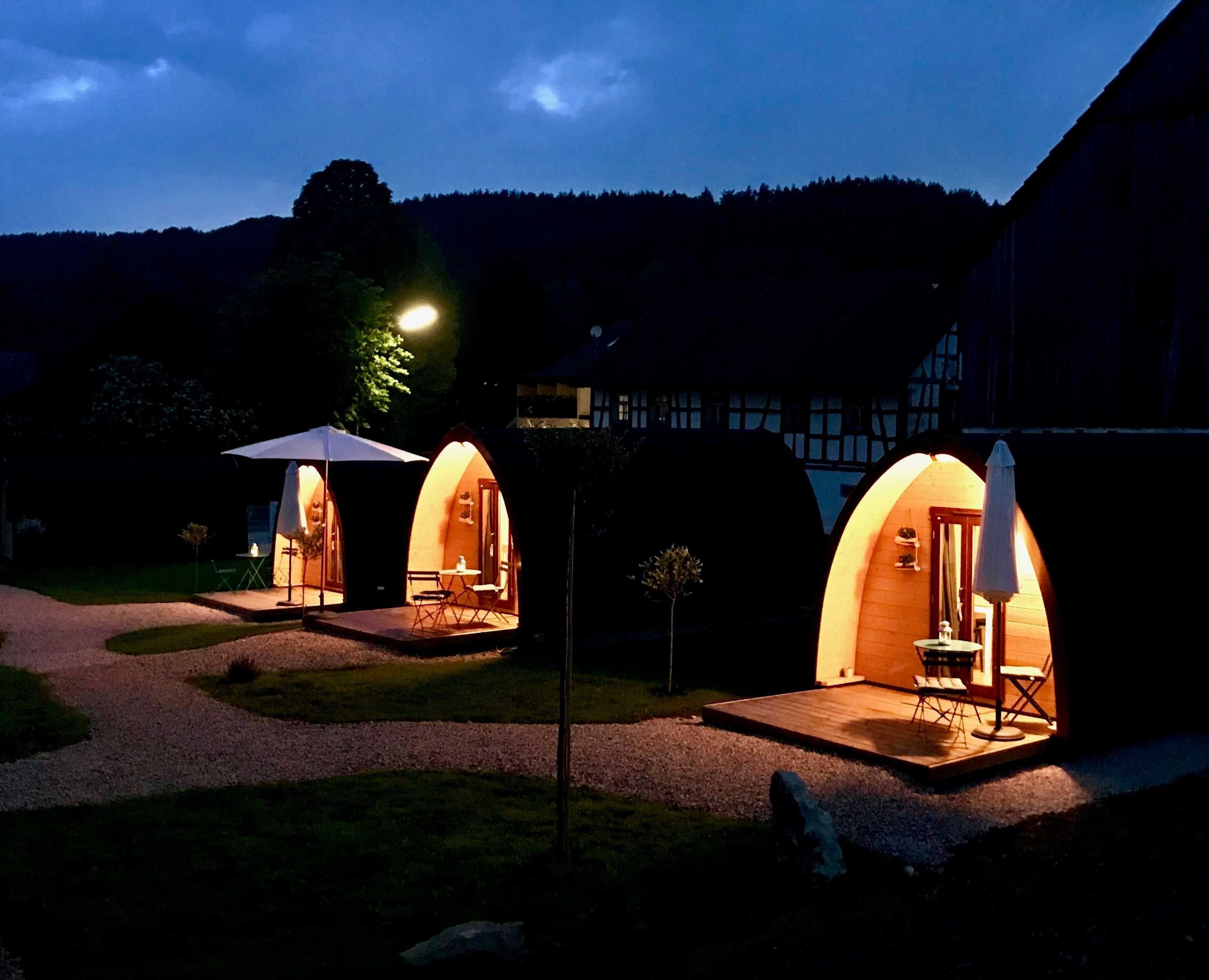 Drie gewelfde, verlichte buiteneeenheden voor overnachting met zitplaatsen en tafels, gelegen voor een gebouw en beboste heuvels.