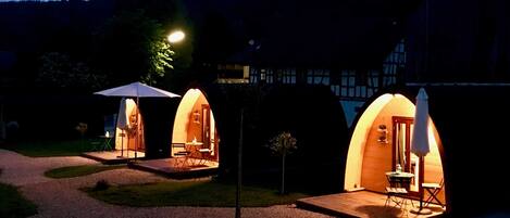 Three arched, illuminated outdoor accommodation units with seating and tables, set against a building and forested hills.