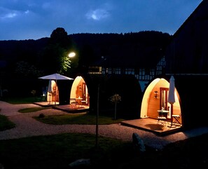 Trois unités d’hébergement extérieures en forme d’arche, illuminées, dotées de sièges et de tables, et situées devant un bâtiment et sur un coteau boisé.