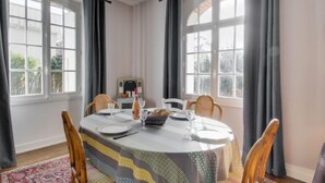 A dining area with a round table set for four, surrounded by chairs. There are two windows with curtains, a geometric light fixture above the table, and a patterned rug on the floor.