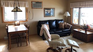 A living room with a dining area, a black leather sofa, a white armchair, a ceiling fan, and a window with a view. - Lake Miltona, MN Weekly Rental Cabin is located right on the beach. (Miltona)