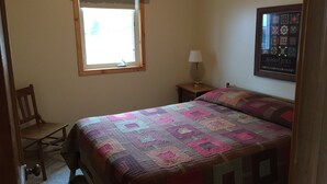 A bedroom with a bed, a chair, a nightstand with a lamp, a window with a curtain, and a quilted wall hanging. - Lake Miltona, MN Weekly Rental Cabin is located right on the beach. (Miltona)