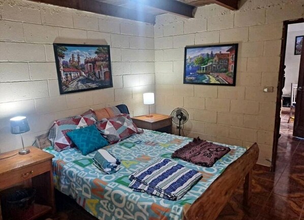 A bedroom with a bed, a nightstand, a lamp, a fan, and two paintings on the wall.