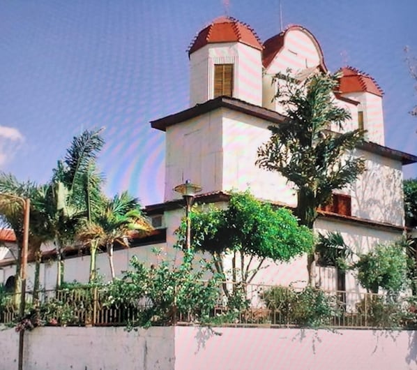 A two-story building with a red-tiled roof and a white facade, surrounded by greenery.