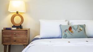 Un lit bien fait, avec du linge de lit blanc et un coussin décoratif, une table de chevet en bois avec une lampe, et un livre posé sur la table.