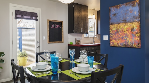 Un coin repas avec une table ronde dressée pour quatre personnes, entourée de chaises. On aperçoit une fenêtre avec des stores, une porte sur laquelle est affiché un panneau « Bienvenue », et un espace cuisine en arrière-plan.
