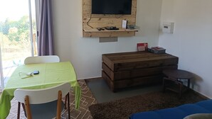 A room with a television mounted on a wooden panel, a table with a green tablecloth, a wooden chest of drawers, and a small round table. - DOMAINE DECOTTE BUNGALOW in the heart of nature (Sainte-Suzanne)