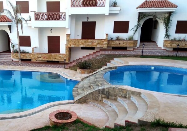 A pool area with two swimming pools, stone steps, and a building with balconies and windows.