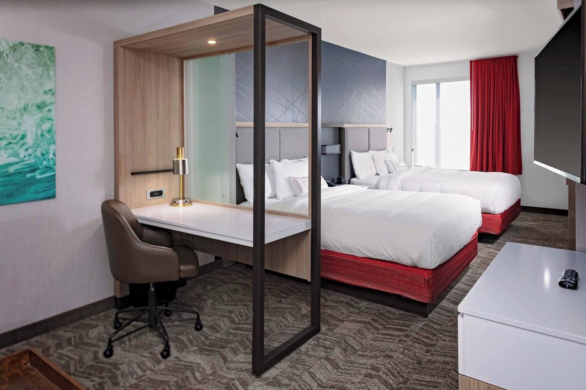 A hotel room with a desk, two beds, a TV, and a window with a red curtain.