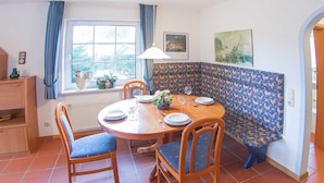 Un coin repas comprenant une table ronde en bois, deux chaises, un banc avec un dossier à motifs et une fenêtre avec des rideaux.