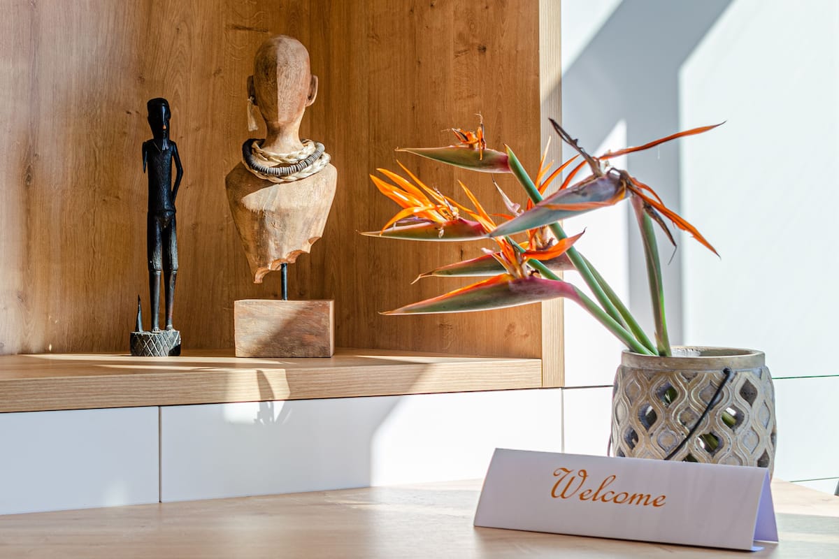 Een ontvangsthal met een houten receptiebalie, een welkomstbord, een borstbeeld en een vogelbekbloem in een decoratieve pot.