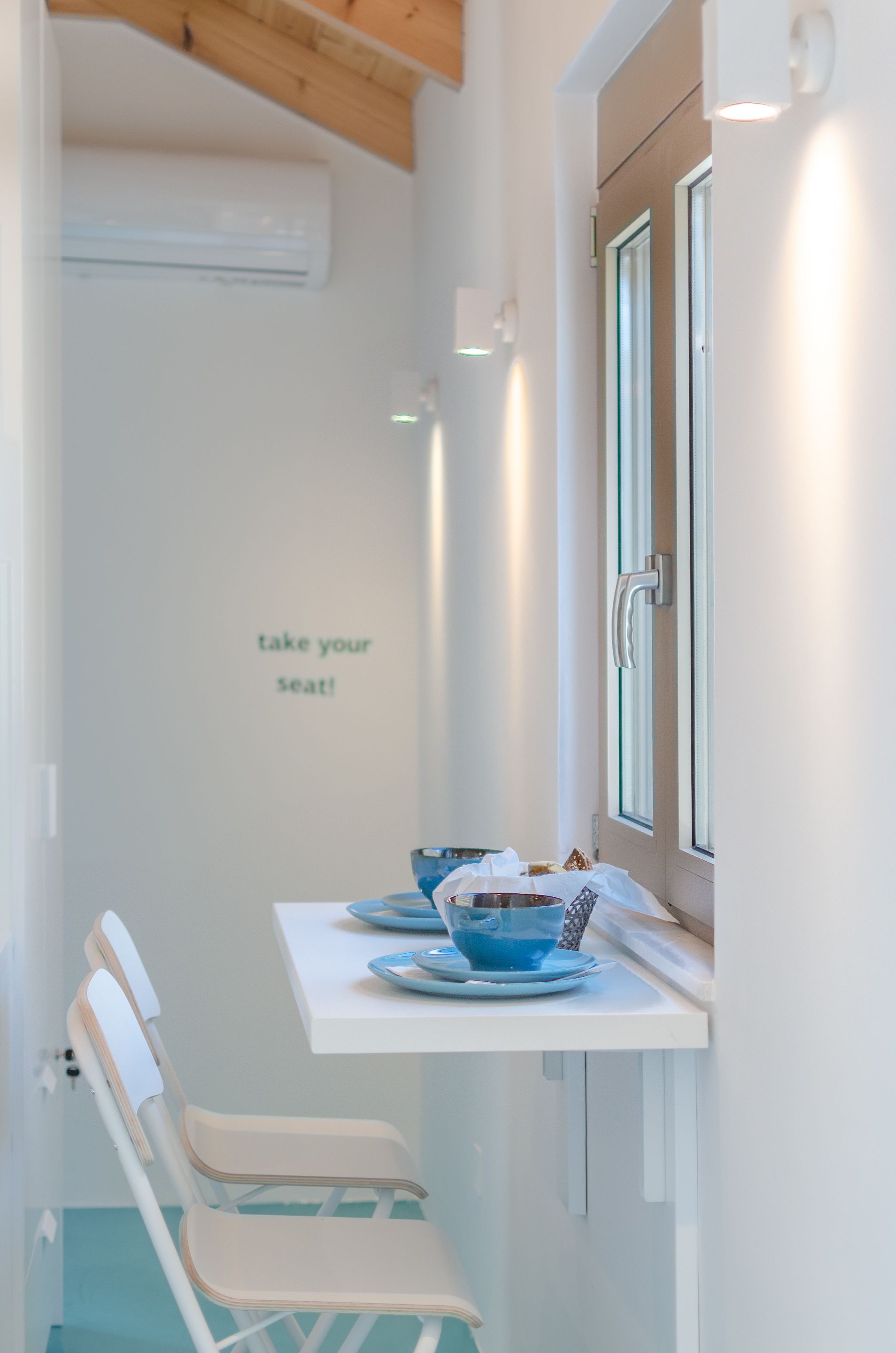 A narrow corridor with a white wall, a door on the right, and a sign that says "take your seat!" A small table with a blue bowl and plates is set up against the wall, and there are two chairs with white and teal cushions.