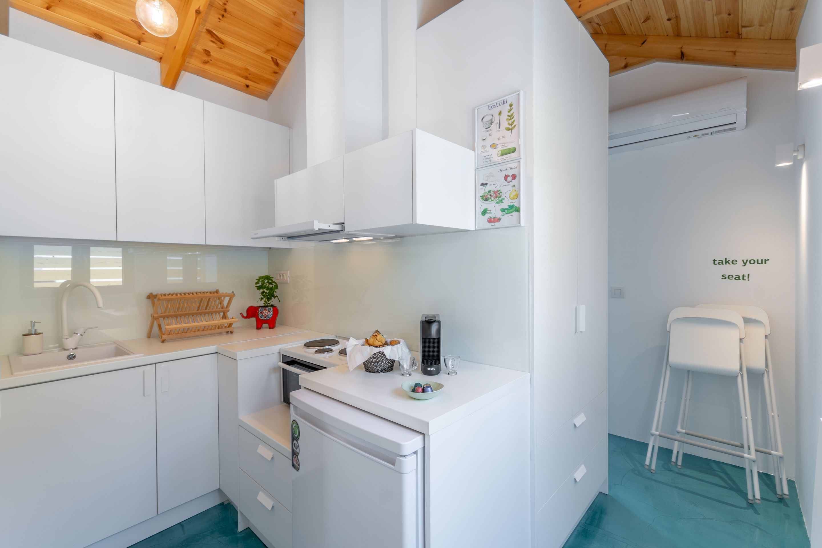 Une cuisine moderne avec des armoires blanches, un four encastré et une machine à café. Le plafond est en bois et un climatiseur est fixé au mur.