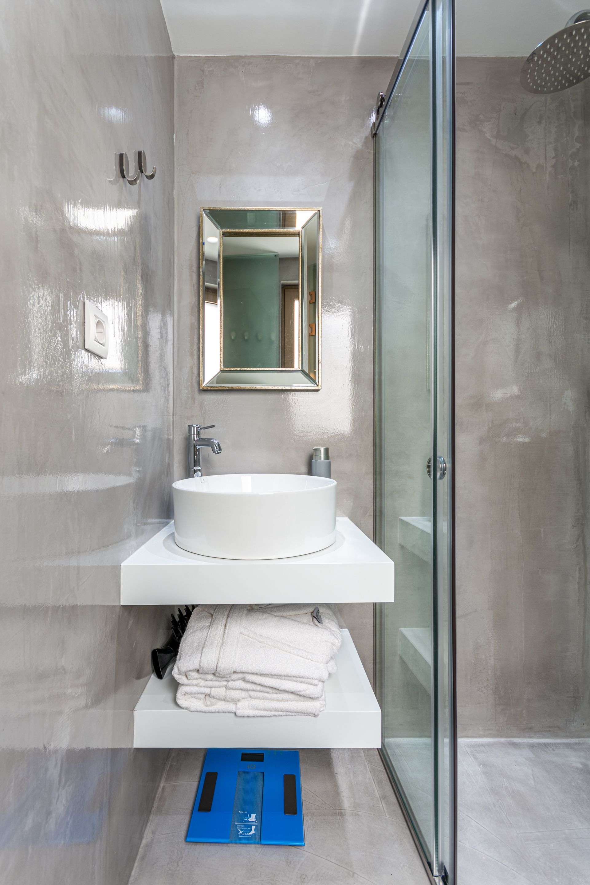 Ein modernes Badezimmer mit einer begehbaren Glasdusche, einem weißen Waschbecken mit goldener Spiegel und einer blauen Waage auf dem Boden.