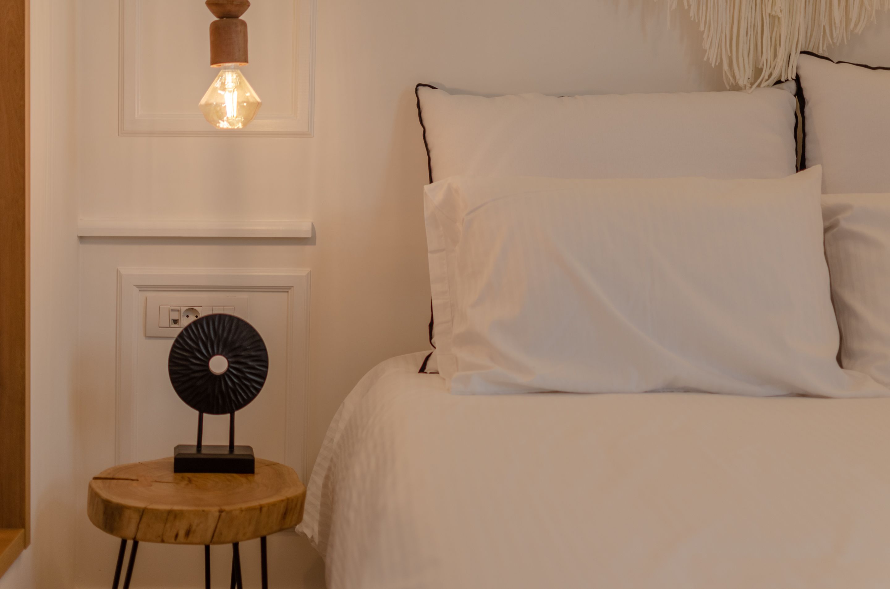 Una cama bien hecha con sábanas blancas, una mesita redonda de madera y un objeto decorativo negro, además de una lámpara de pared.