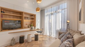 A modern living room with a large flat-screen TV mounted on the wall, wooden shelves, a herringbone-patterned floor, a sofa, and a dining area with a table and chairs.