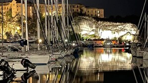 Una marina di notte con barche ormeggiate, un edificio imponente sullo sfondo e una scogliera rocciosa.