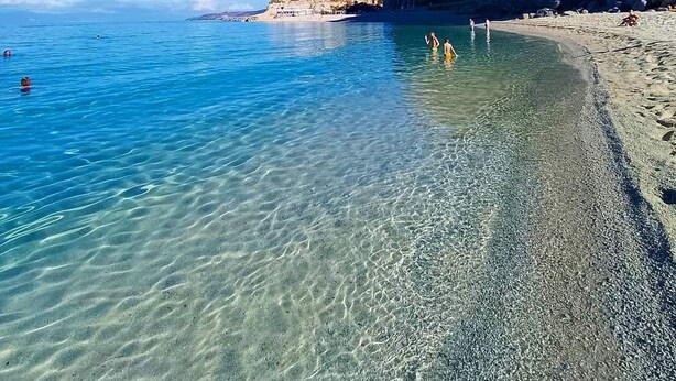 Una spiaggia assolata con acque calme, una scogliera rocciosa e alcune persone in lontananza.