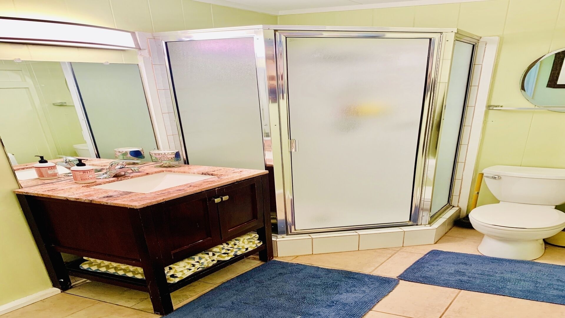 A bathroom with a vanity, sink, mirror, shower enclosure, toilet, and blue rugs.