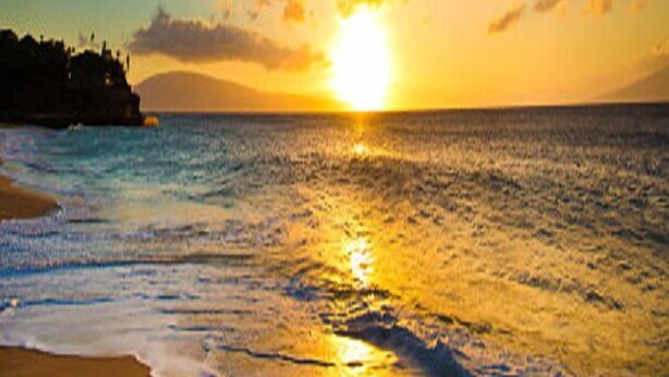 Una playa con el sol poniéndose sobre el océano, creando una silueta de tierra a lo lejos.