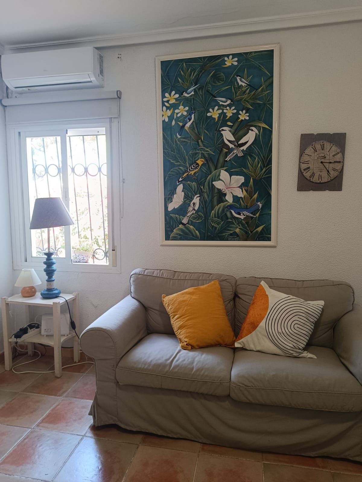 Un salon avec un canapé beige, une lampe, une horloge murale et un tableau représentant un oiseau tropical.