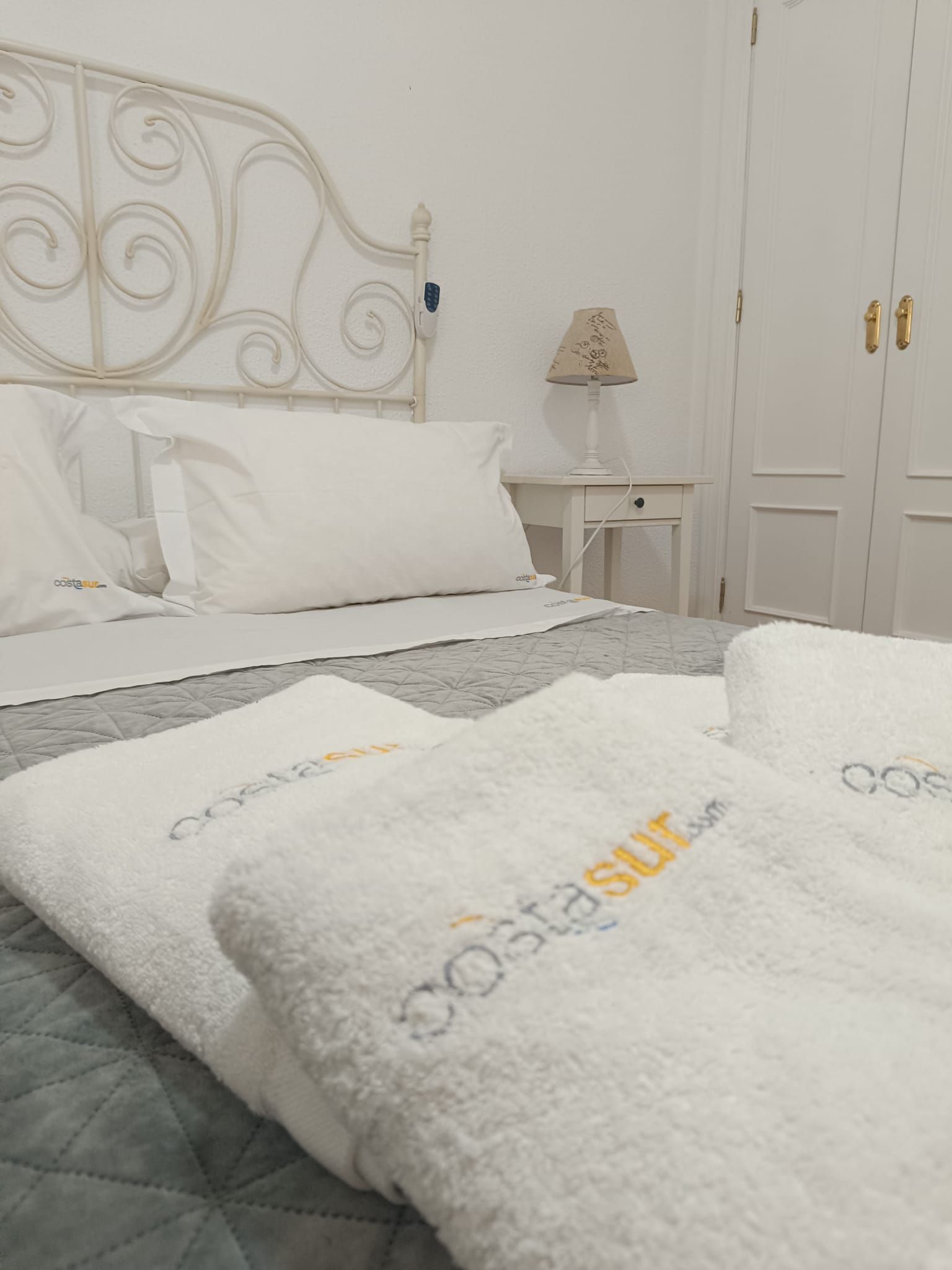 A neatly made bed with white linens and a grey quilt, a bedside table with a lamp, and a wardrobe with double doors.