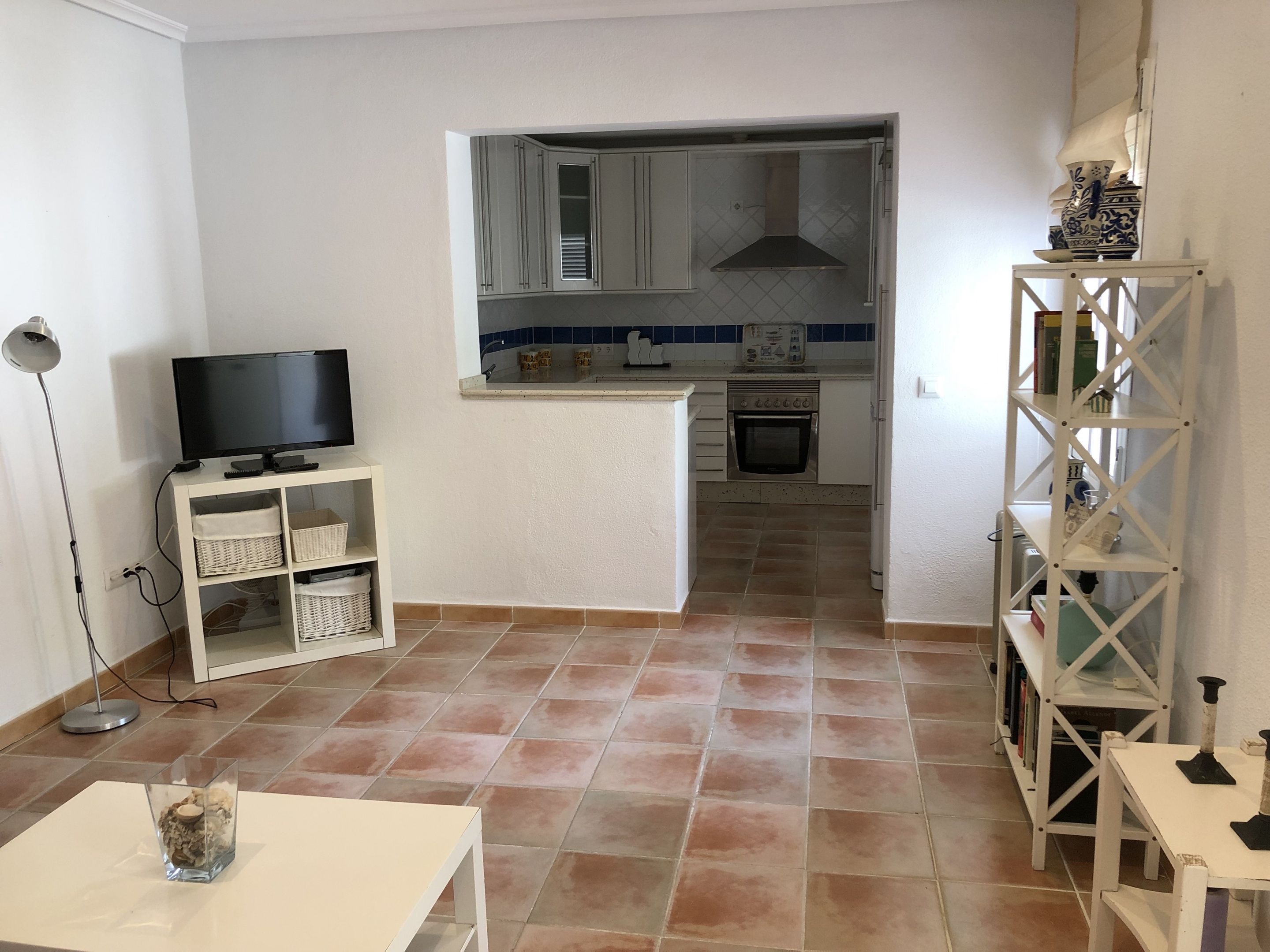 Un espacio habitable pequeño con una televisión sobre un mueble bajo, una mesita y una zona de cocina con armarios y una estufa.