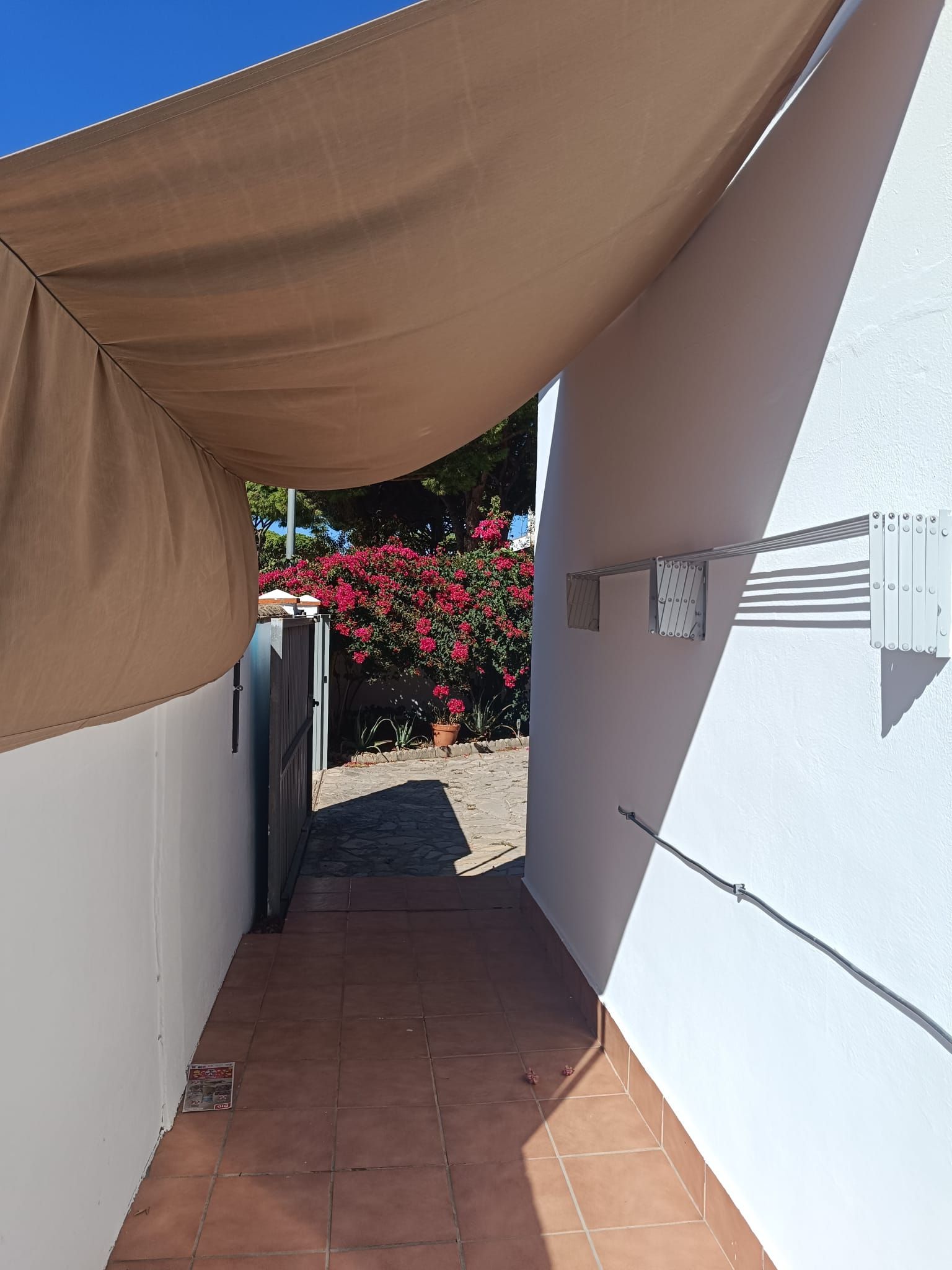 A narrow outdoor corridor with a brown canopy, a white wall, and a potted plant with red flowers.