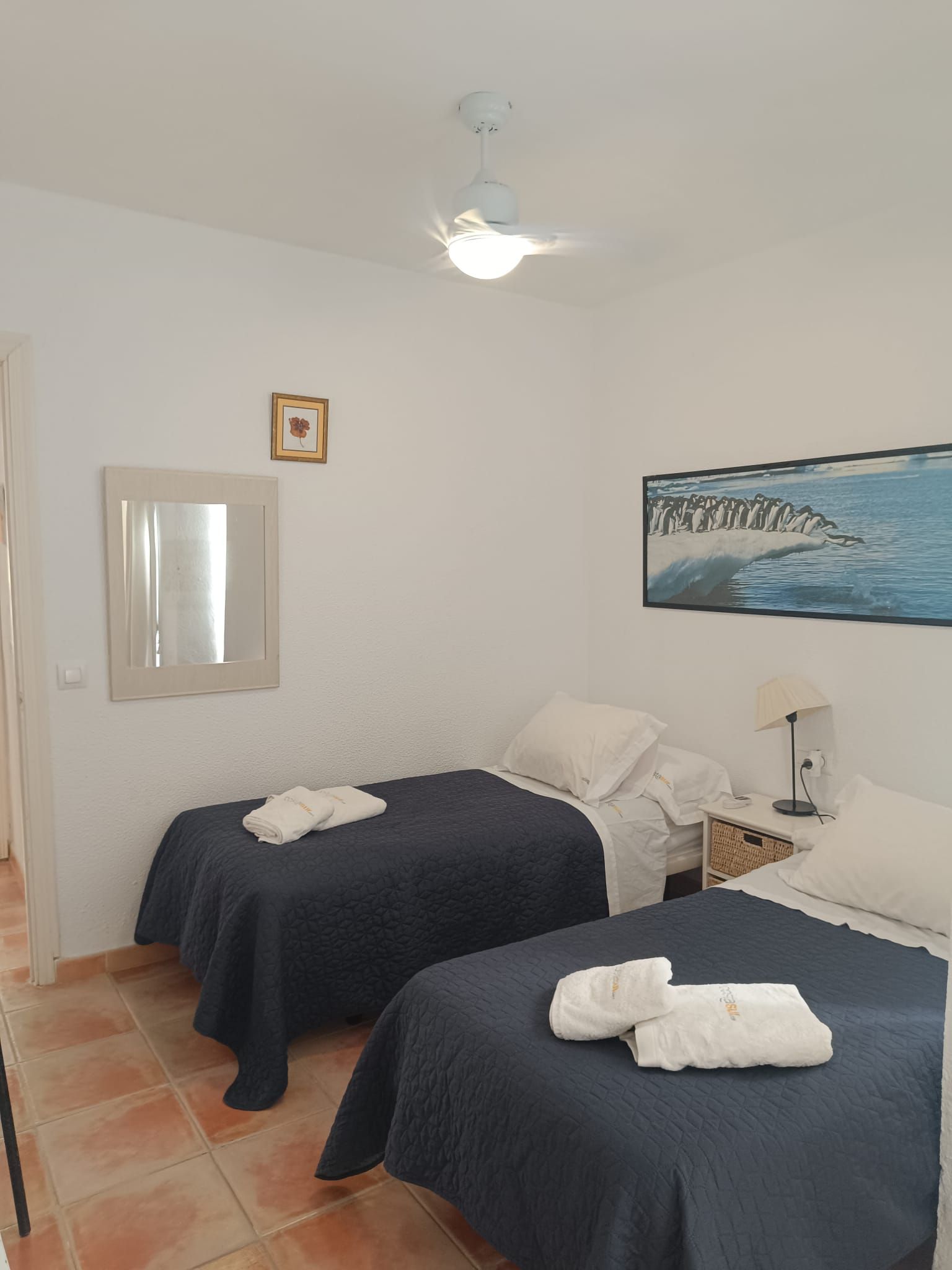 A hotel room with two beds, a ceiling fan, a framed picture, and a painting of penguins.