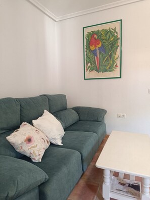 A green sofa with floral cushions, a white table, and a framed botanical illustration on the wall.