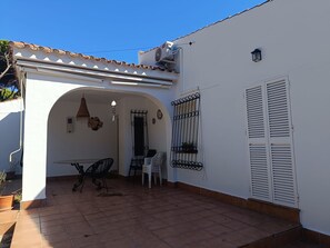 A patio with a table and chairs, a white wall with a window, and a door.