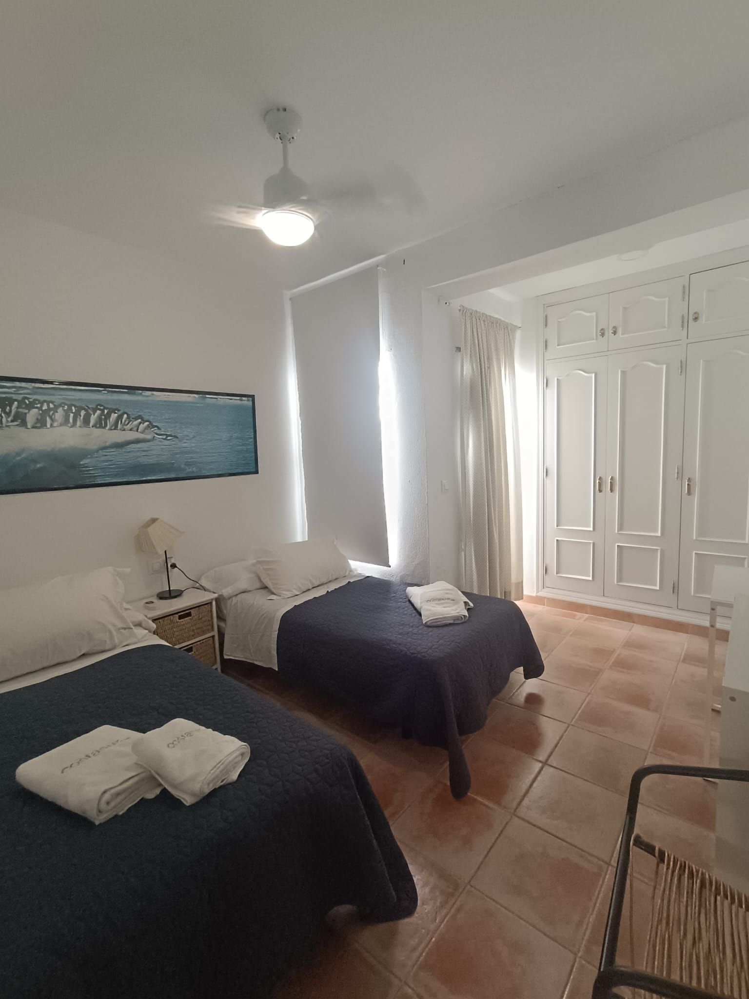 A room with two beds, a ceiling fan, a painting of penguins, and a wardrobe.