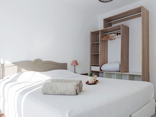 A neatly made bed with a white comforter and a rolled towel, a bedside table with a lamp and a cup, and a large mirror with a wooden frame and shelves.