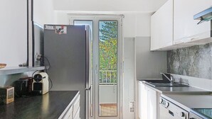 A modern kitchen with white cabinets, a stainless steel refrigerator, a built-in oven, and a large window overlooking a balcony with greenery. - Le Clos tranquille by Interhome (Nice)