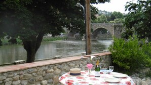 Ein Steintisch mit einer Tischdecke mit Blumenmuster, eine Champagnerflasche und Gläser auf einer Flussufer-Terrasse mit einer Steinmauer und einer Brücke im Hintergrund.