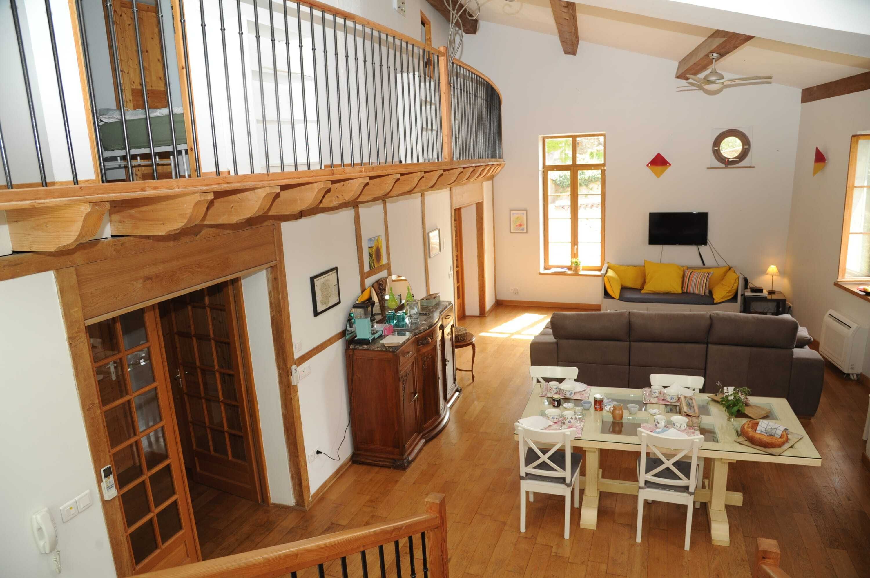 A spacious living area with a high ceiling, wooden balcony railing, a sofa, a dining table with chairs, and a television.