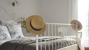 Een bed met een grijze deken, een wit metalen kinderbedje, en een stroohoed op het bedje.