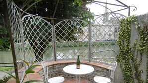 Een terras met twee witte metalen stoelen en een tafel, een fles wijn, en zicht op een rivier en gebouwen op de achtergrond.