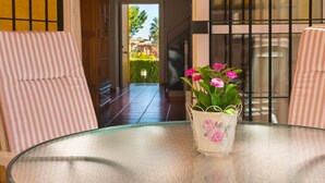 A glass table with a white vase containing pink flowers, two striped chairs, and a view of a garden through a glass door. - Cozy Townhouse in Islantilla: Enjoy the Beach, Golf and Adventures (Lepe)