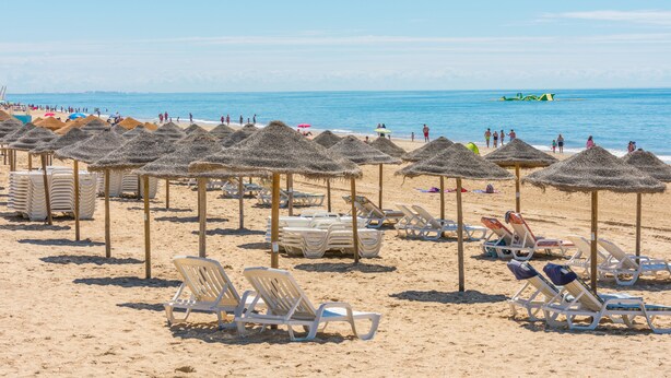 A beach with rows of sun loungers and thatched umbrellas, a sandy shore, and the ocean in the background. - Cozy Townhouse in Islantilla: Enjoy the Beach, Golf and Adventures (Lepe)