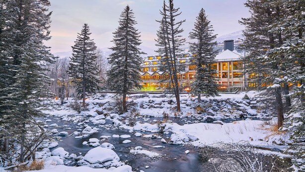 A lodge with a river in the foreground, surrounded by snow-covered trees. - Spacious Condo within walking distance to Chair 20 & access  to Grand Hyatt Amenities (Vail)