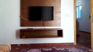 A room with a mounted TV on a wooden panel, a lower shelf, and a patterned rug on the floor. - Residence Alcazaba Oued Laou 4129 (OUED LAW)